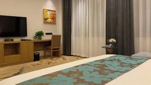 A hotel room with a bed, a desk, a chair, a television, and a painting on the wall. - Jinjiaxiang Hotel (Xiji)