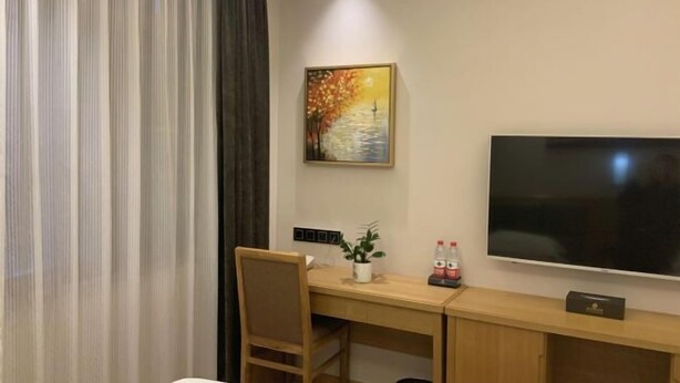 A hotel room with a wooden desk, a chair, a flat-screen TV, a painting, and a potted plant. - Jinjiaxiang Hotel (Xiji)