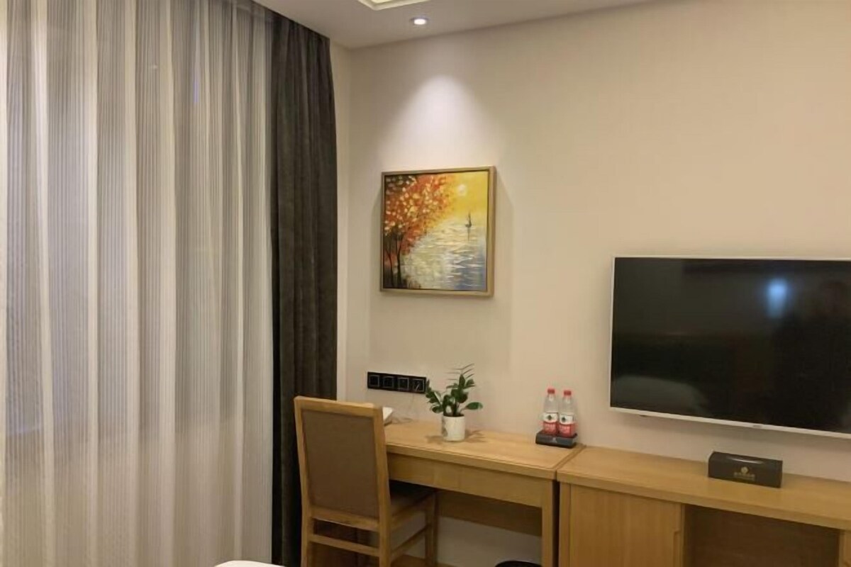 A hotel room with a wooden desk, a chair, a flat-screen TV, a painting, and a potted plant.