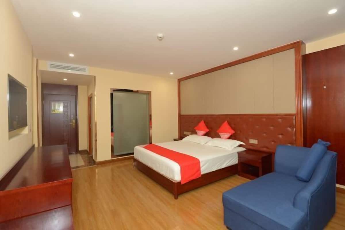 A hotel room with a bed, a blue armchair, a wooden desk, and a TV on the wall.
