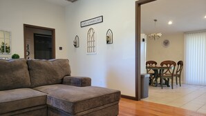 A living room with a sectional sofa, a dining area with a table and chairs, and a 'Live a Good Life' sign on the wall.