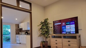 A modern living room with a flat-screen TV displaying a streaming service interface, a wooden cabinet, a potted plant, and a view into the kitchen.
