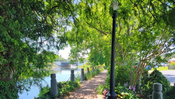 A brick pathway lined with greenery and a lamp post, leading to a body of water with a bridge in the background.