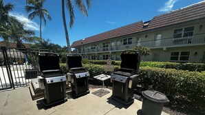A patio with three barbecue grills, a trash can, and a small table. - 1BR Marco Island condo with pool, WiFi, kitchen, and heated pool. (Marco Island)