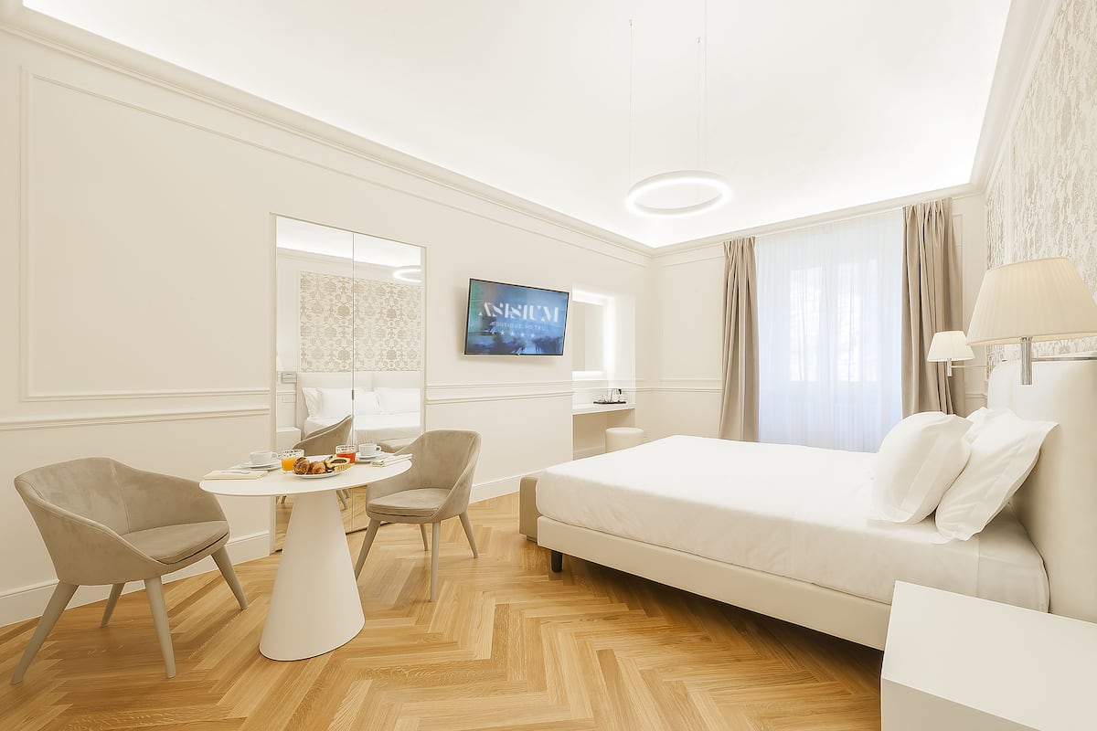 A hotel room with a bed, two chairs, a small table with a drink, a TV, and a window with curtains.