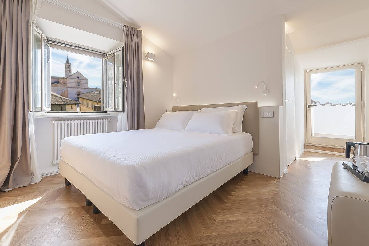A neatly arranged bedroom with a large bed, a window with curtains, a radiator, and a door leading to another room.