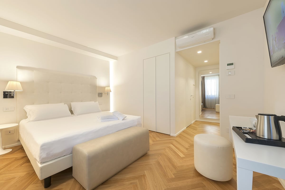 A modern hotel room with a large bed, bedside tables, a desk with a kettle, and a TV.