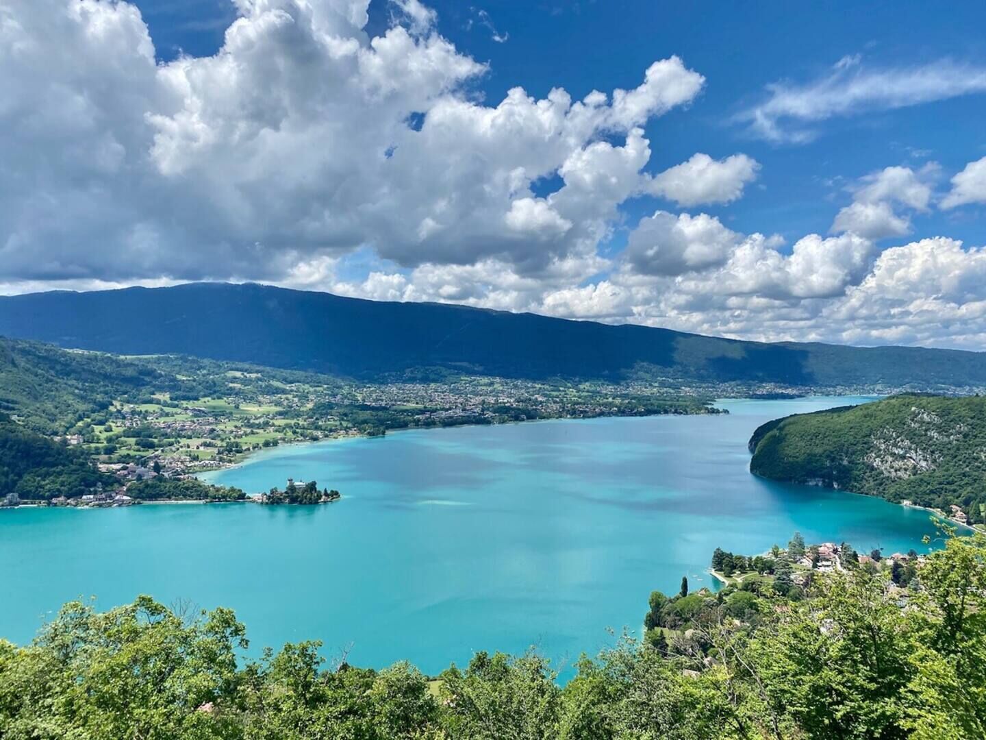 Vista panorâmica de uma cidade costeira com uma grande baía turquesa, rodeada por colinas e montanhas verdejantes.