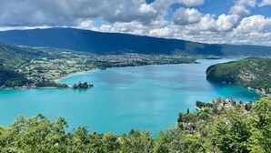 A panoramic view of a coastal town with a large turquoise bay, surrounded by lush green hills and mountains. - The Bergerie T3- view Mountains. (Talloires-Montmin)