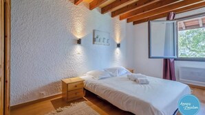 A bedroom with a bed, nightstands, a wall-mounted light, a window with a view, and a ceiling with wooden beams. - The Bergerie T3- view Mountains. (Talloires-Montmin)