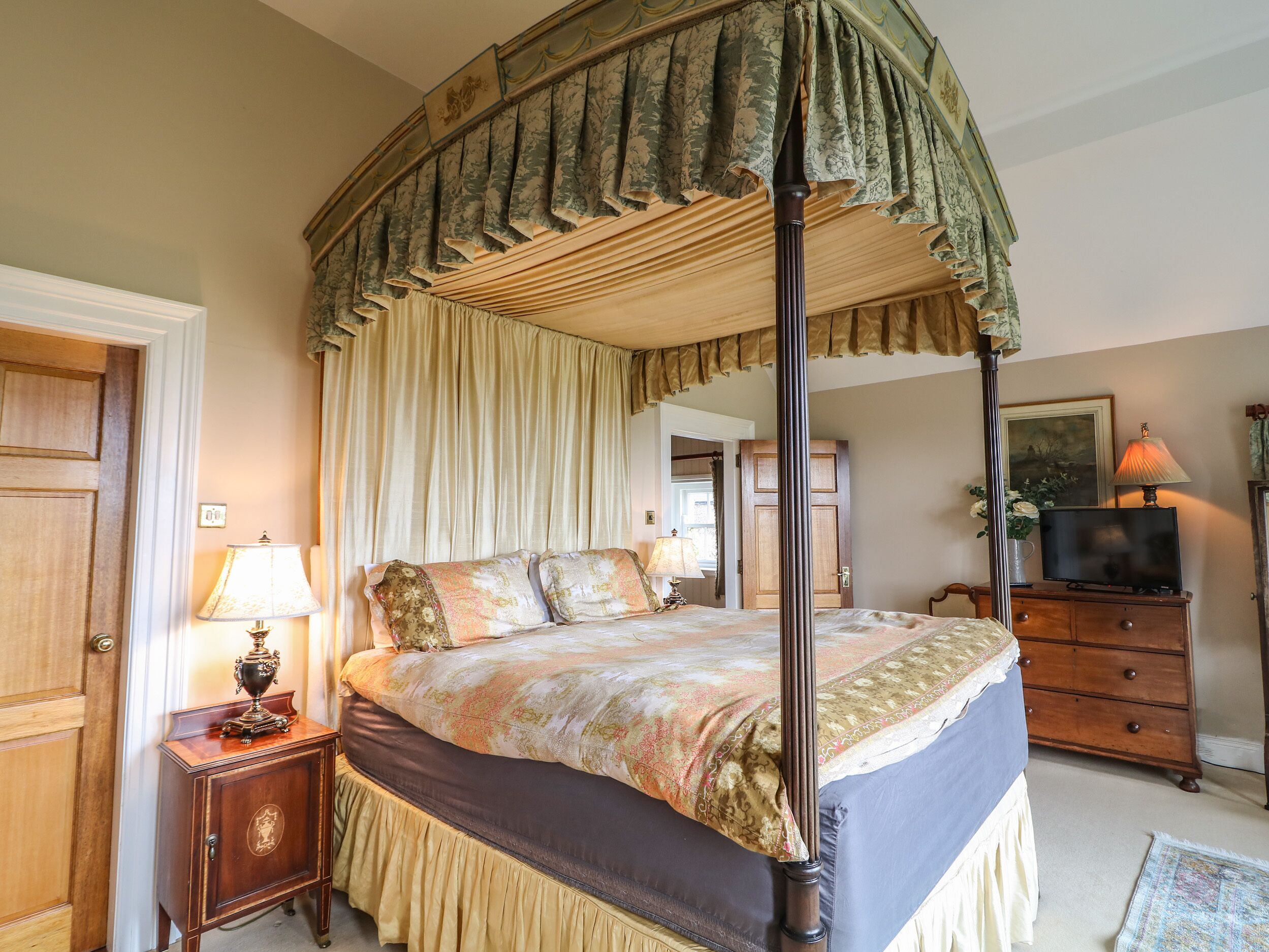 A four-poster canopy bed with a matching canopy, a wooden nightstand with a lamp, a dresser with a television, and a door with a brass handle.
