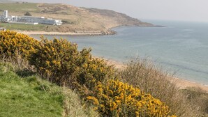 Un paysage côtier avec un ensemble de bâtiments situé près du rivage, offrant une vue dégagée sur la mer et les collines lointaines.