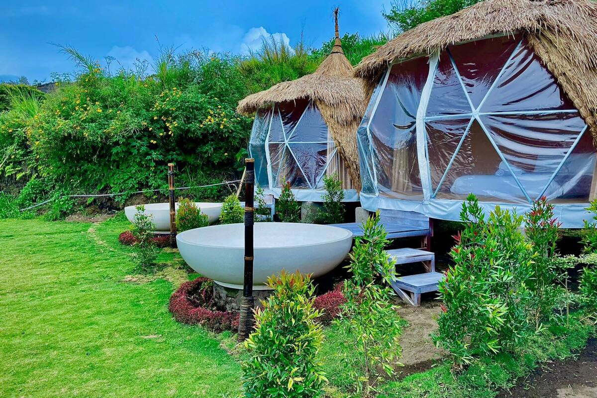 A garden with thatched-roof structures, a large white basin, and lush greenery.