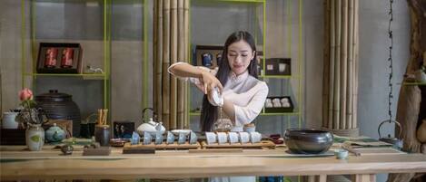 Une personne dispose des tasses à thé sur une table en bois dans une pièce décorée d’éléments en bambou et dotée d’étagères où sont rangés divers objets.