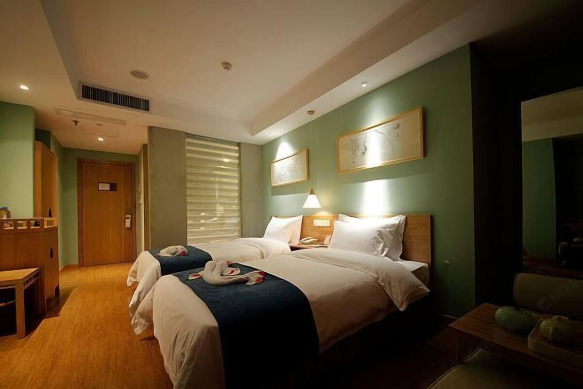 A hotel room with two beds, a desk, a chair, a bedside table, and a large window with blinds.