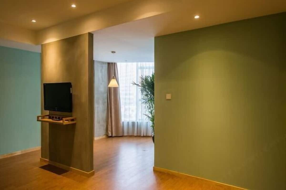 A spacious room with a wooden floor, a flat-screen TV mounted on a wall, and a large window with curtains.