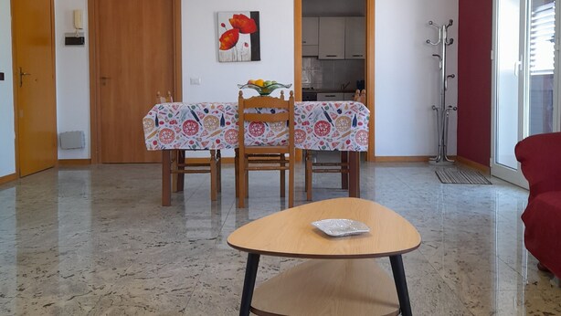 Uma sala ampla com uma mesa de jantar posta para quatro pessoas, uma poltrona vermelha e uma cozinha visível através de uma porta aberta.