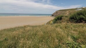 Paisaje costero con playa de arena, zonas verdes y un acantilado con un camino que lleva a un edificio.