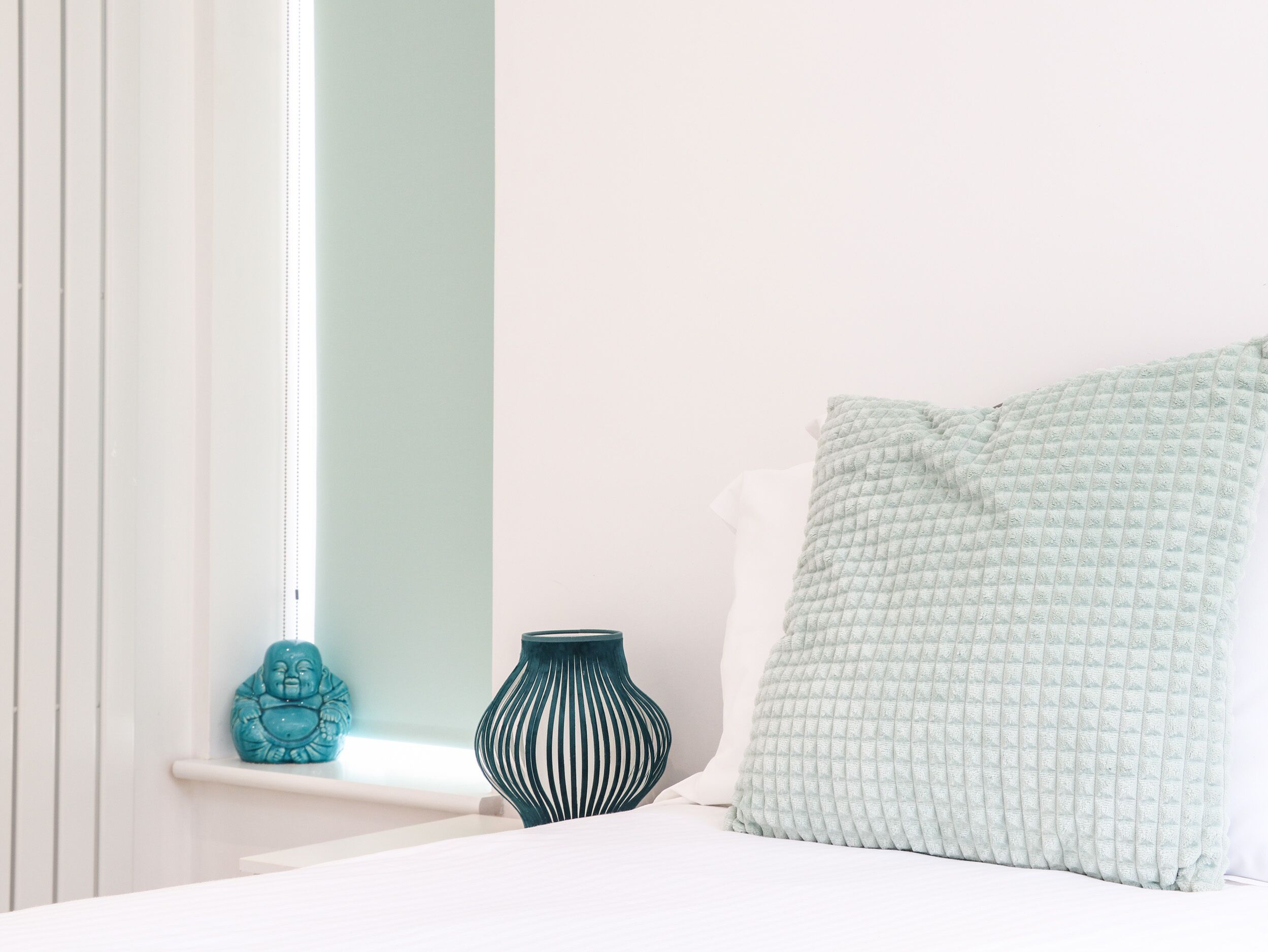 Een slaapkamer met een bed, een raam met zonwering, een decoratief voorwerp in blauwgroen en een vaas.