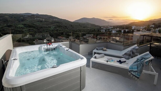 Een dakterras met een jacuzzi, lounge stoelen en een glazen balustrade met uitzicht op een bergachtig landschap tijdens zonsondergang.