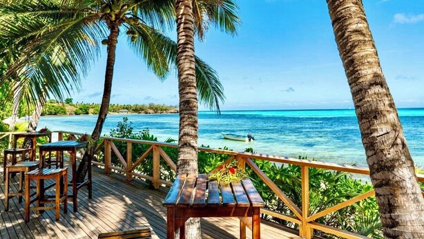 A wooden deck with tables and chairs overlooking a clear blue sea, palm trees, and a distant boat. - Nabua Lodge Hidden Getaway (Nacula Island)