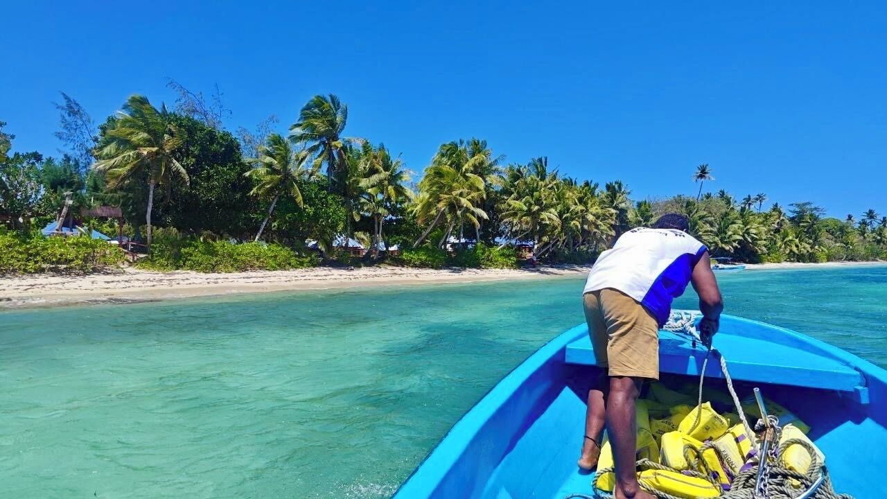 A person in a boat on a clear turquoise sea with a beach and palm trees in the background.