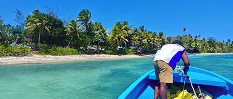 A person in a boat on a clear turquoise sea with a beach and palm trees in the background.
