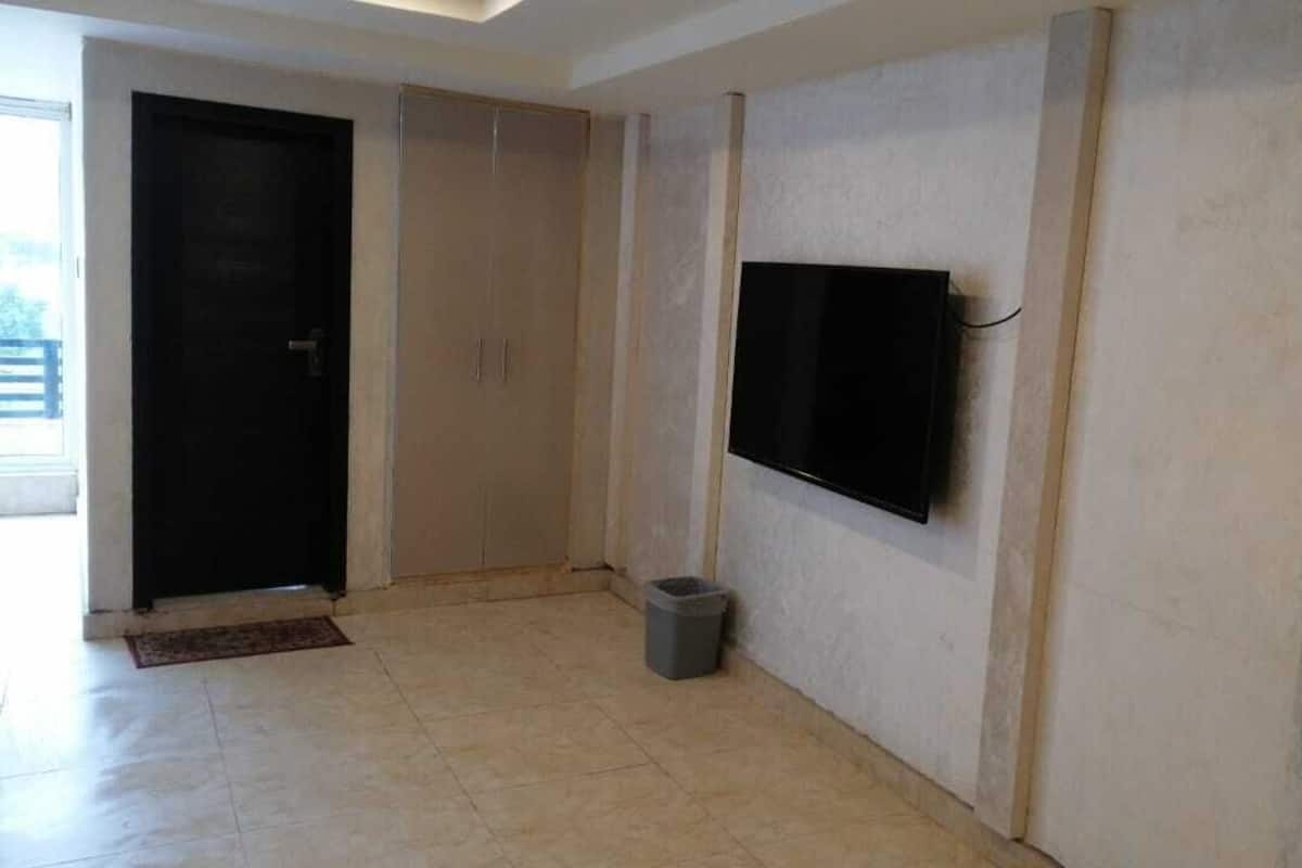 Ein Zimmer mit Fliesenboden, einer schwarzen Tür, einem Schrank, einem an der Wand befestigten Fernseher und einem Mülleimer.