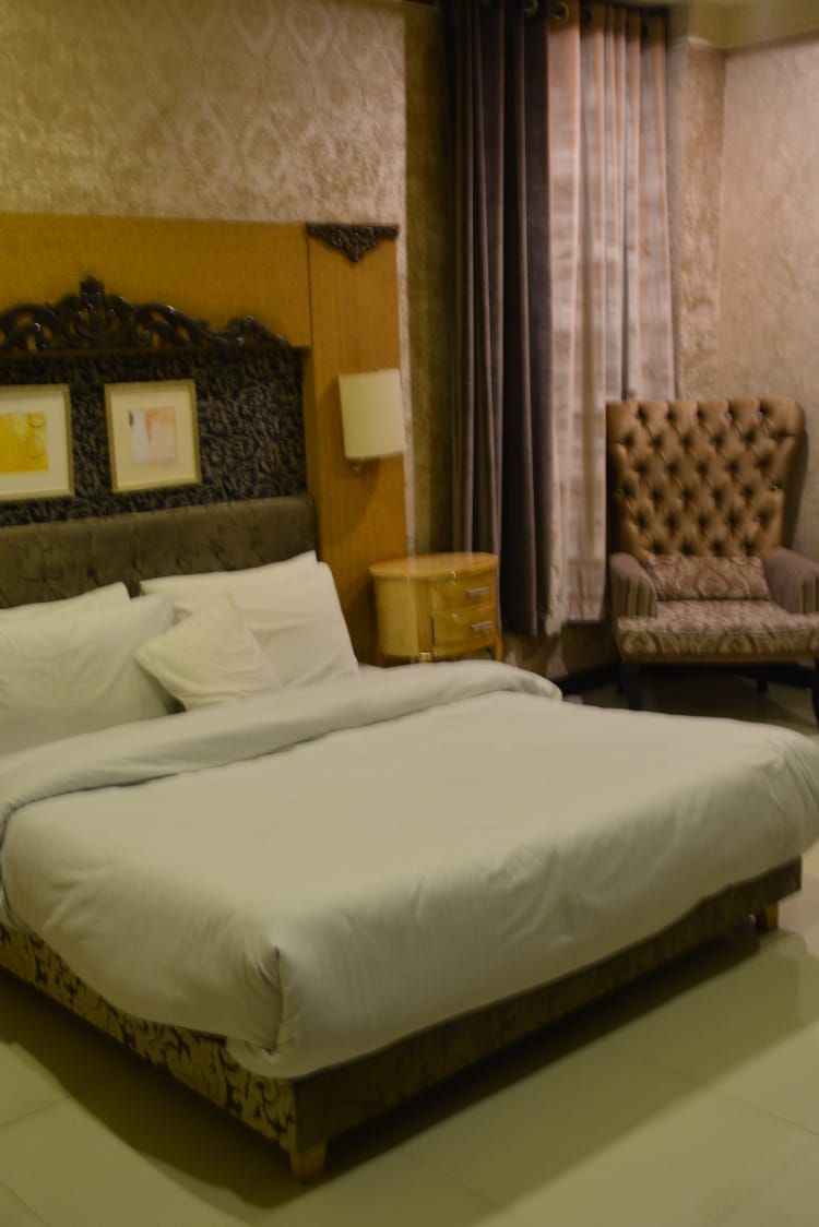 Ein Hotelzimmer mit einem großen Bett, einem hölzernen Kopfteil, einem kleinen Nachttisch, einem Stuhl und einer Wandlampe.