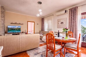 Una sala de estar moderna con un televisor de pantalla plana, un sofá, una mesa de comedor con sillas y un amplio ventanal con cortinas.