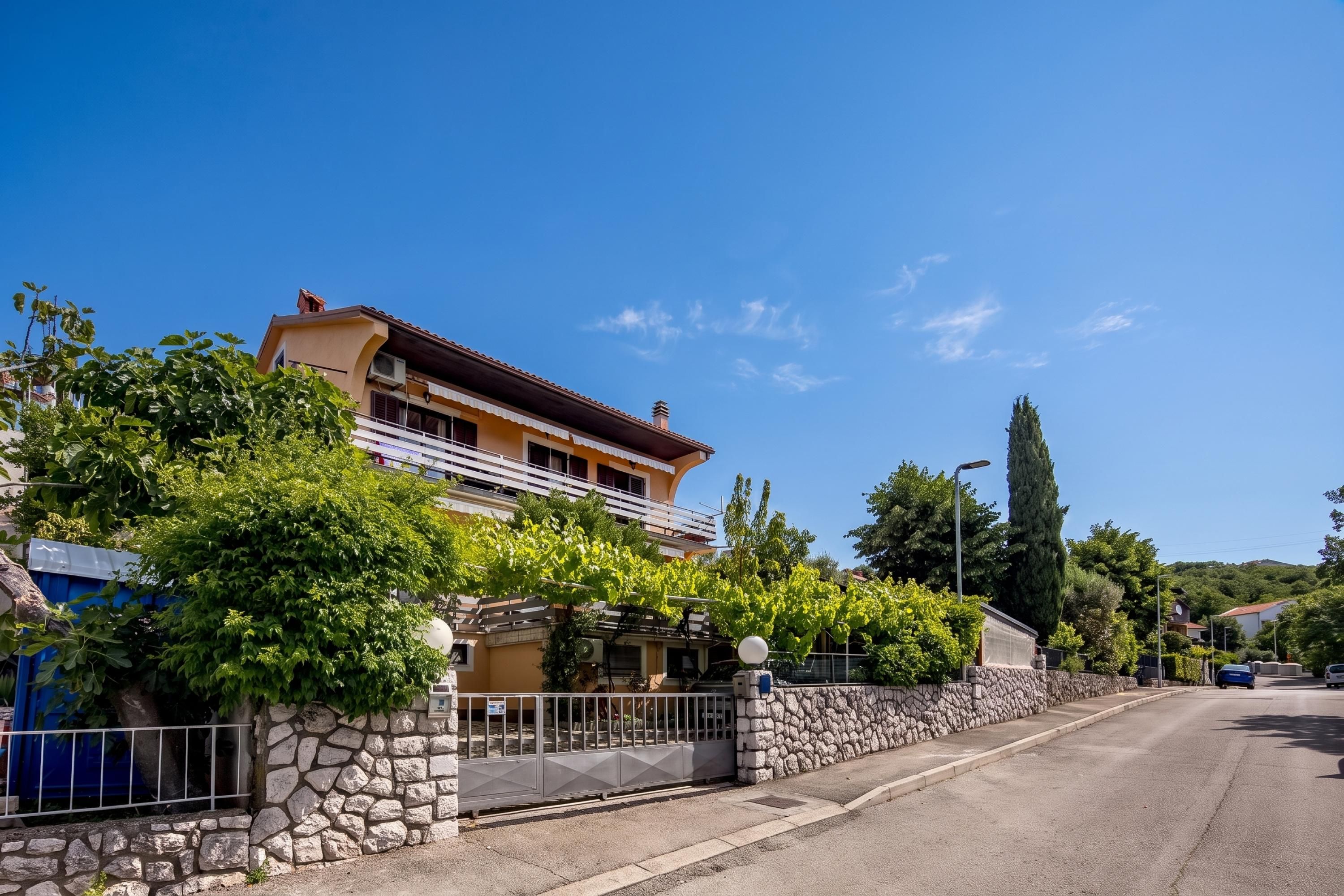 A two-story building with a stone wall and a metal gate, surrounded by greenery and a clear sky.