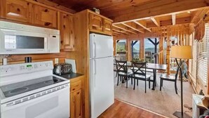 A wooden cabin interior with a kitchen area featuring a stove, microwave, and refrigerator. There is a dining table with chairs and a view of the outdoors through a window. - Majestic 2BD near TOWN! VIEWS + Pool (Pigeon Forge)
