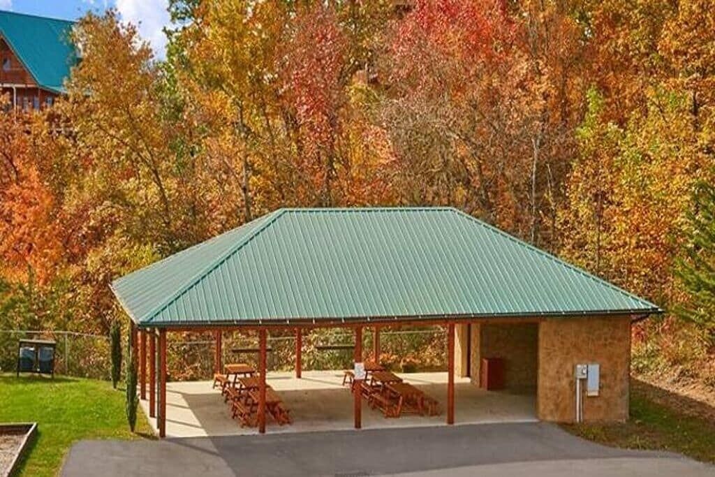 A covered pavilion with a green roof, wooden benches, and a small building adjacent to it.