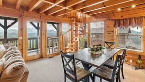 A wooden cabin interior with a dining table, chairs, a staircase, and large windows offering mountain views. - Majestic 2BD near TOWN! VIEWS + Pool (Pigeon Forge)