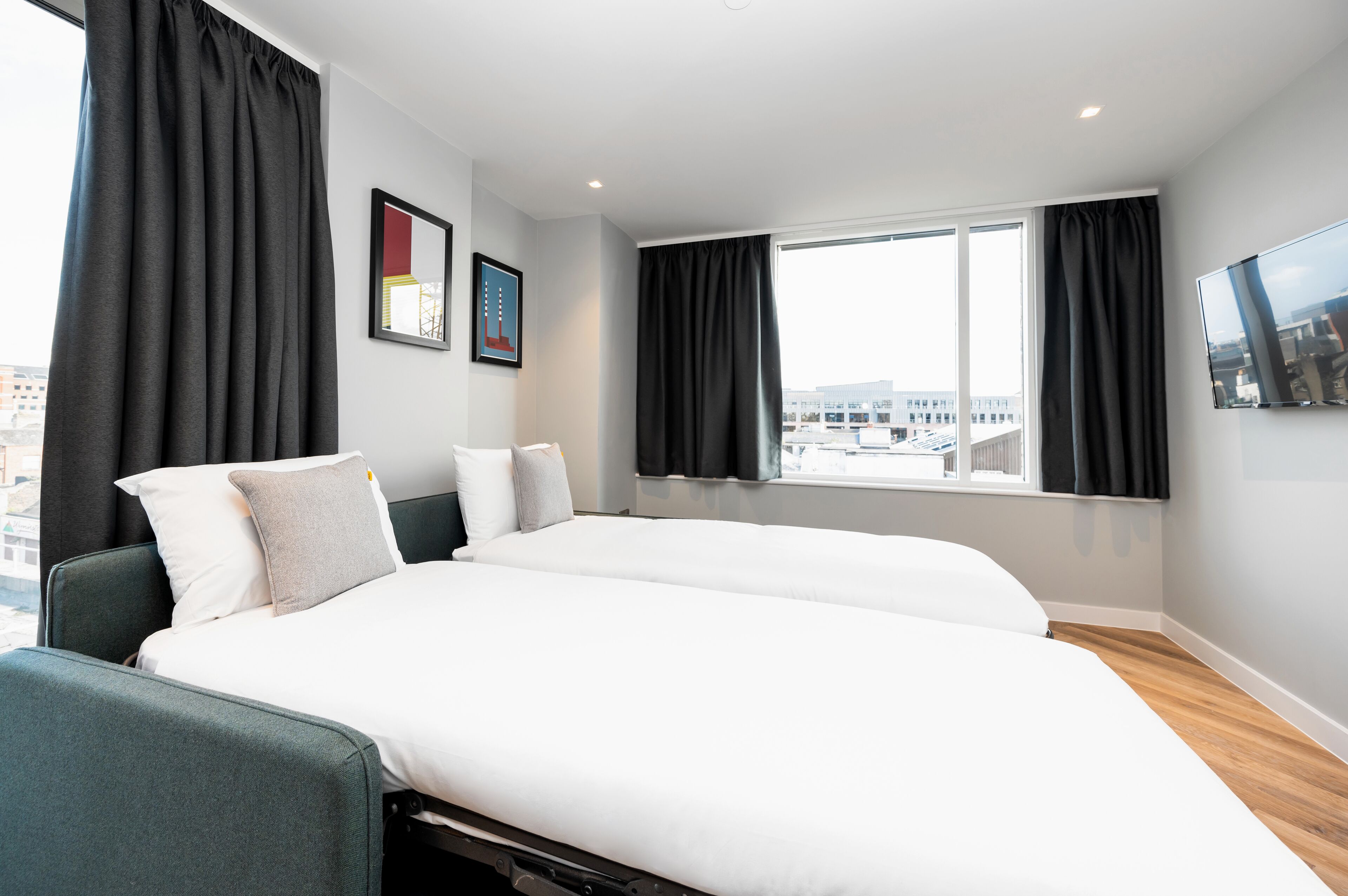 A modern hotel room with two beds, large windows, and a city view.