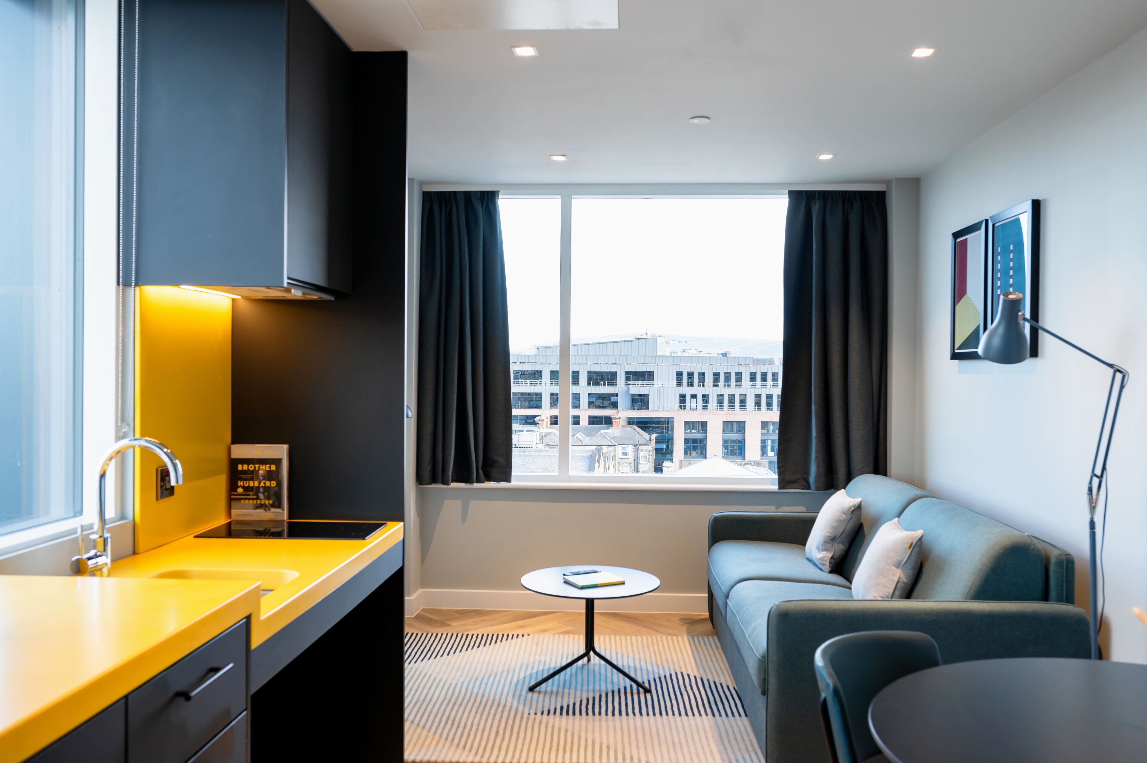 A modern hotel room with a kitchen area, a sofa, a small table, and a large window offering a city view.