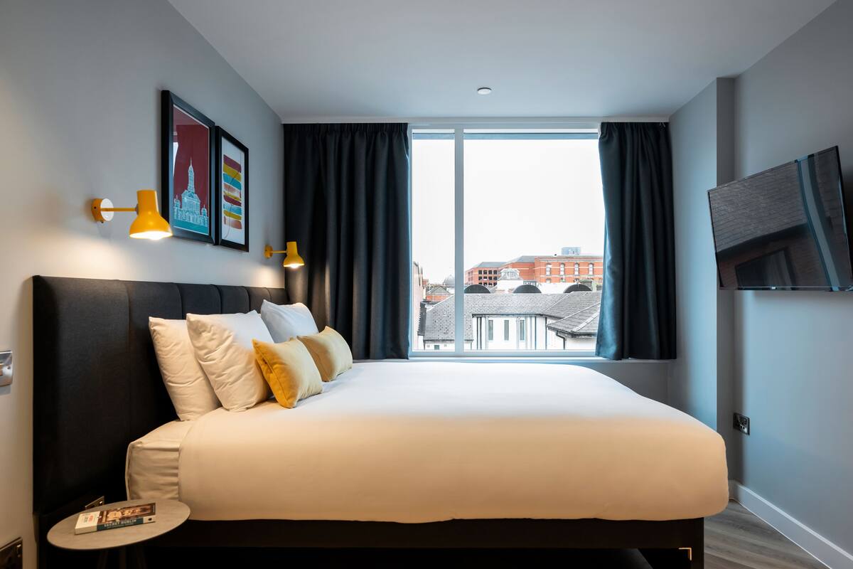 Une chambre d’hôtel moderne avec un grand lit, une petite table de chevet, un téléviseur et une vue sur la ville grâce à de grandes fenêtres.