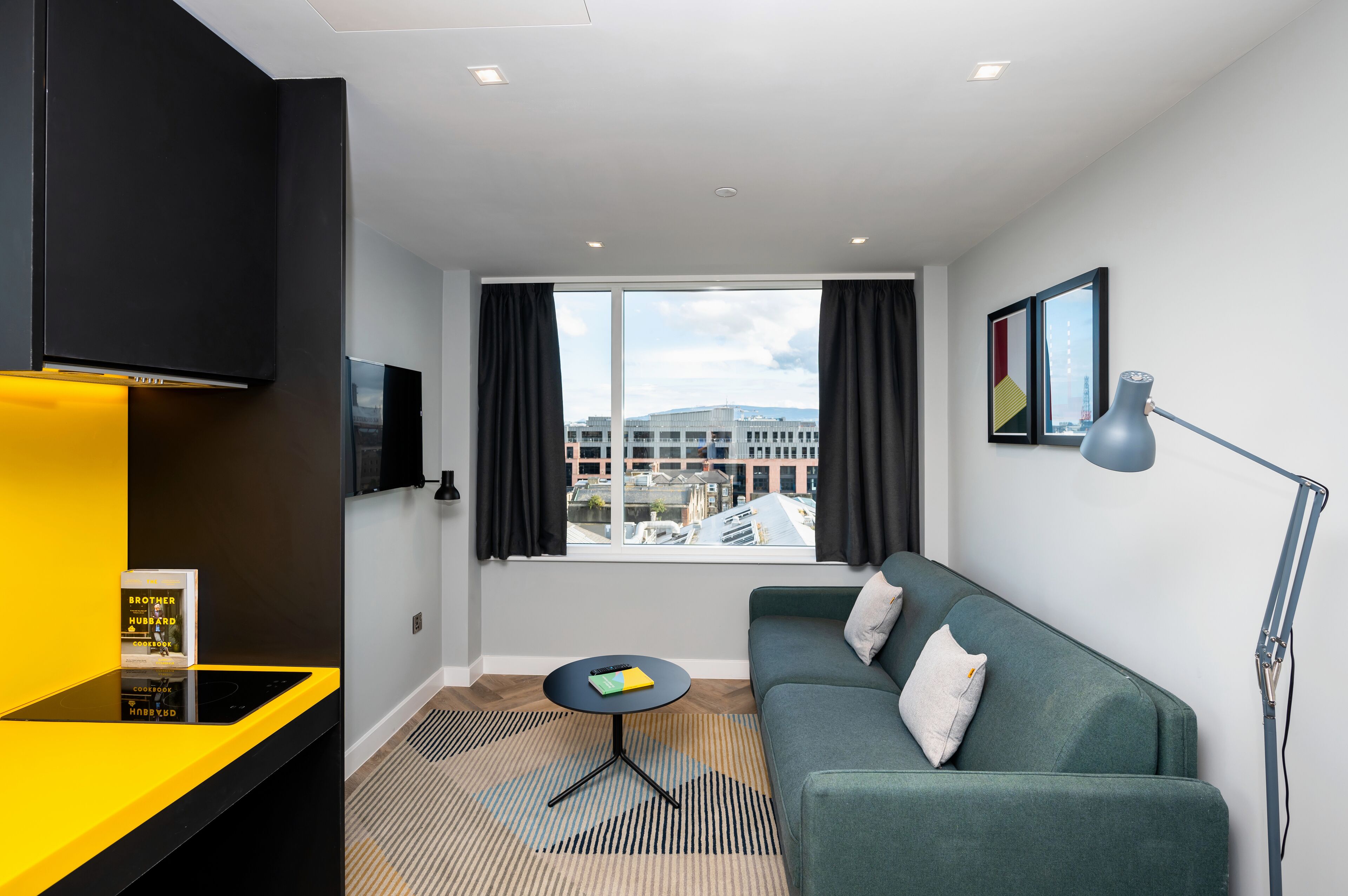 Una habitación de hotel moderna con un sofá verde, una mesita redonda, un televisor negro y una ventana con cortinas.