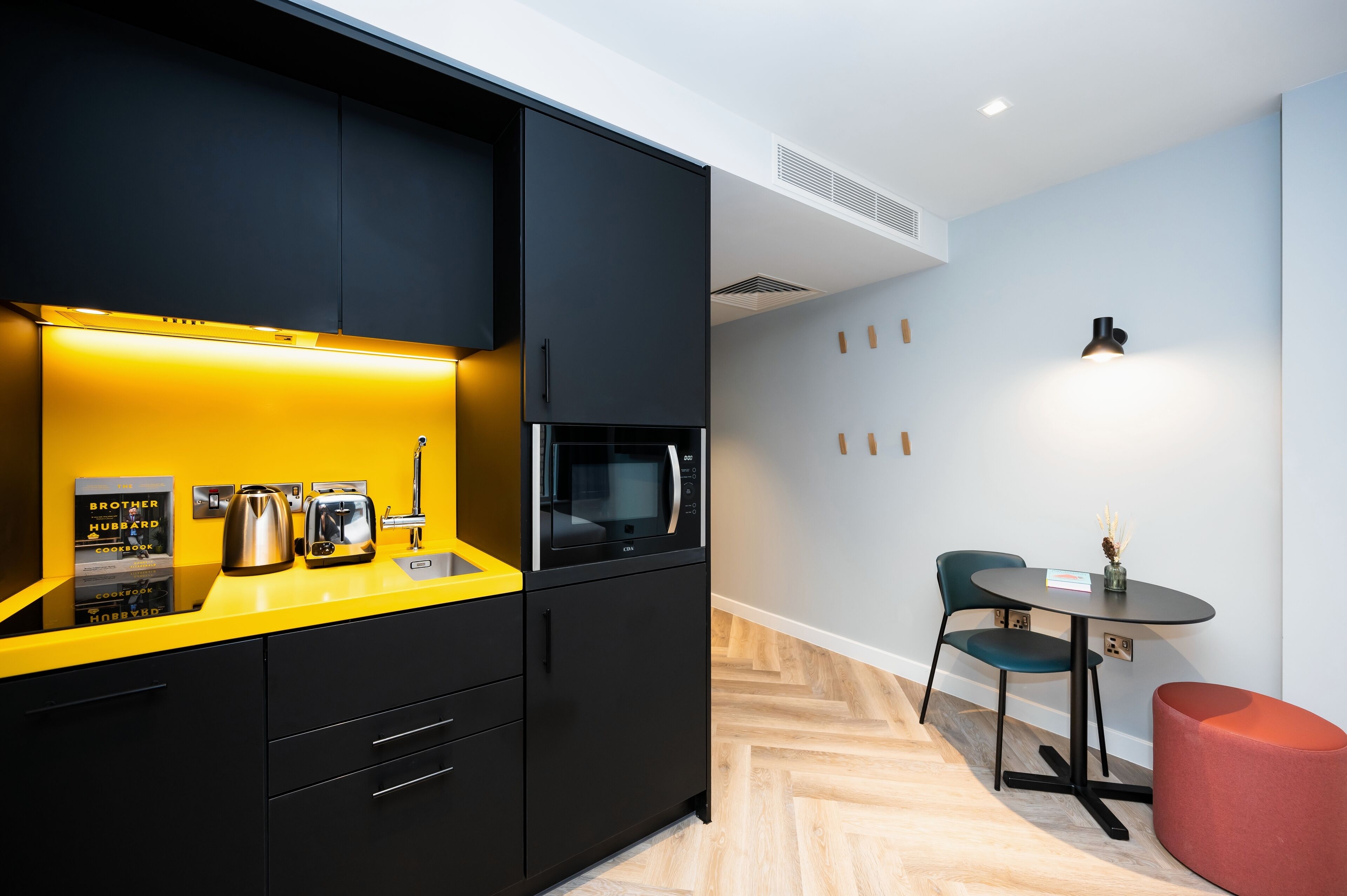 Una cocina moderna con armarios negros, una encimera amarilla y un microondas. Hay una pequeña mesa con una silla y un reposapiés rojo.