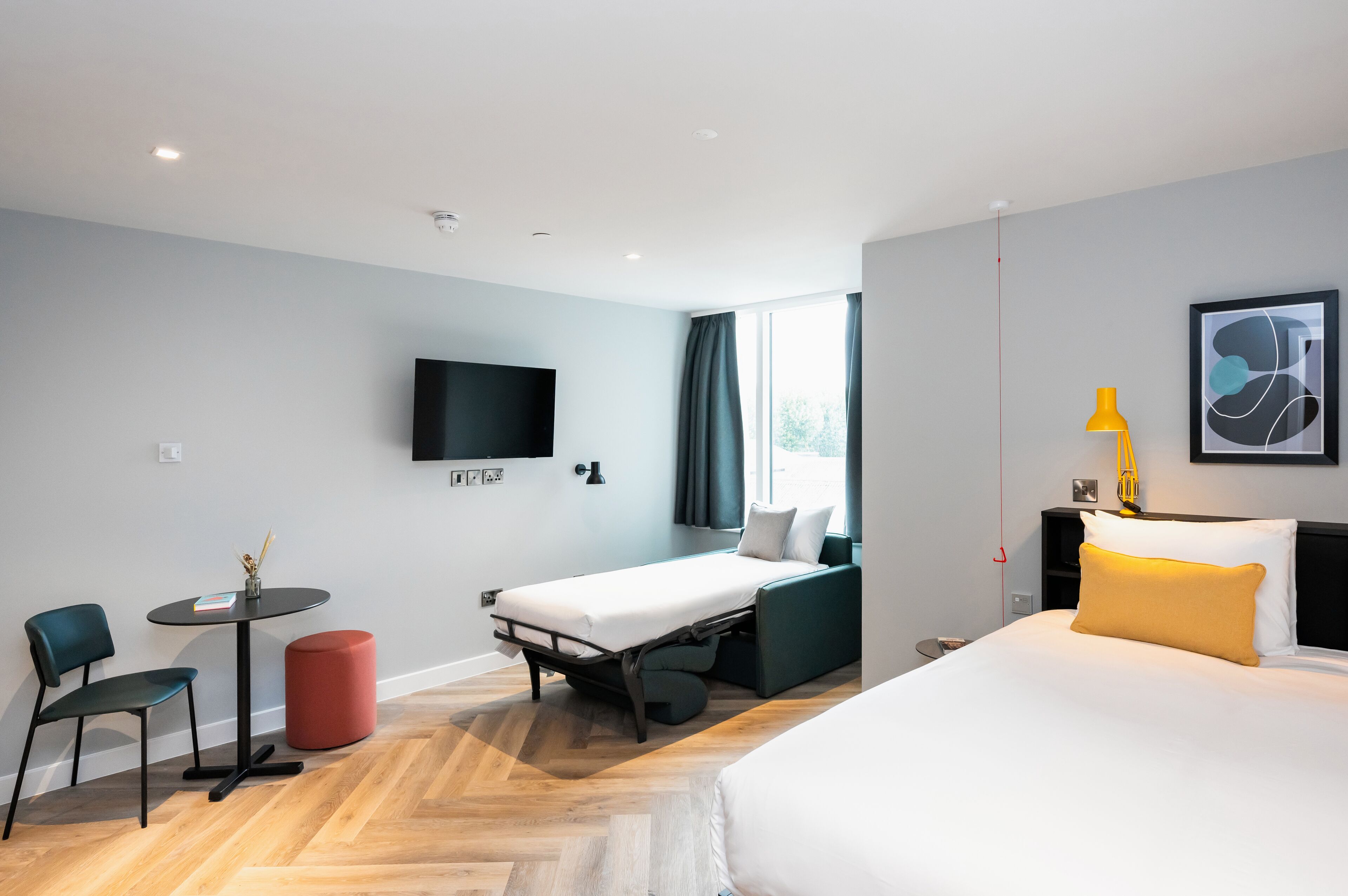 A modern hotel room with a bed, a desk, a chair, a small table, and a TV.