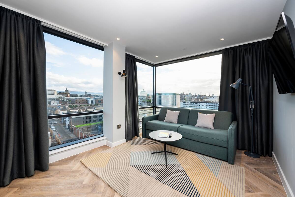 Un salon moderne avec une grande fenêtre, un canapé vert, une petite table et un lampadaire.