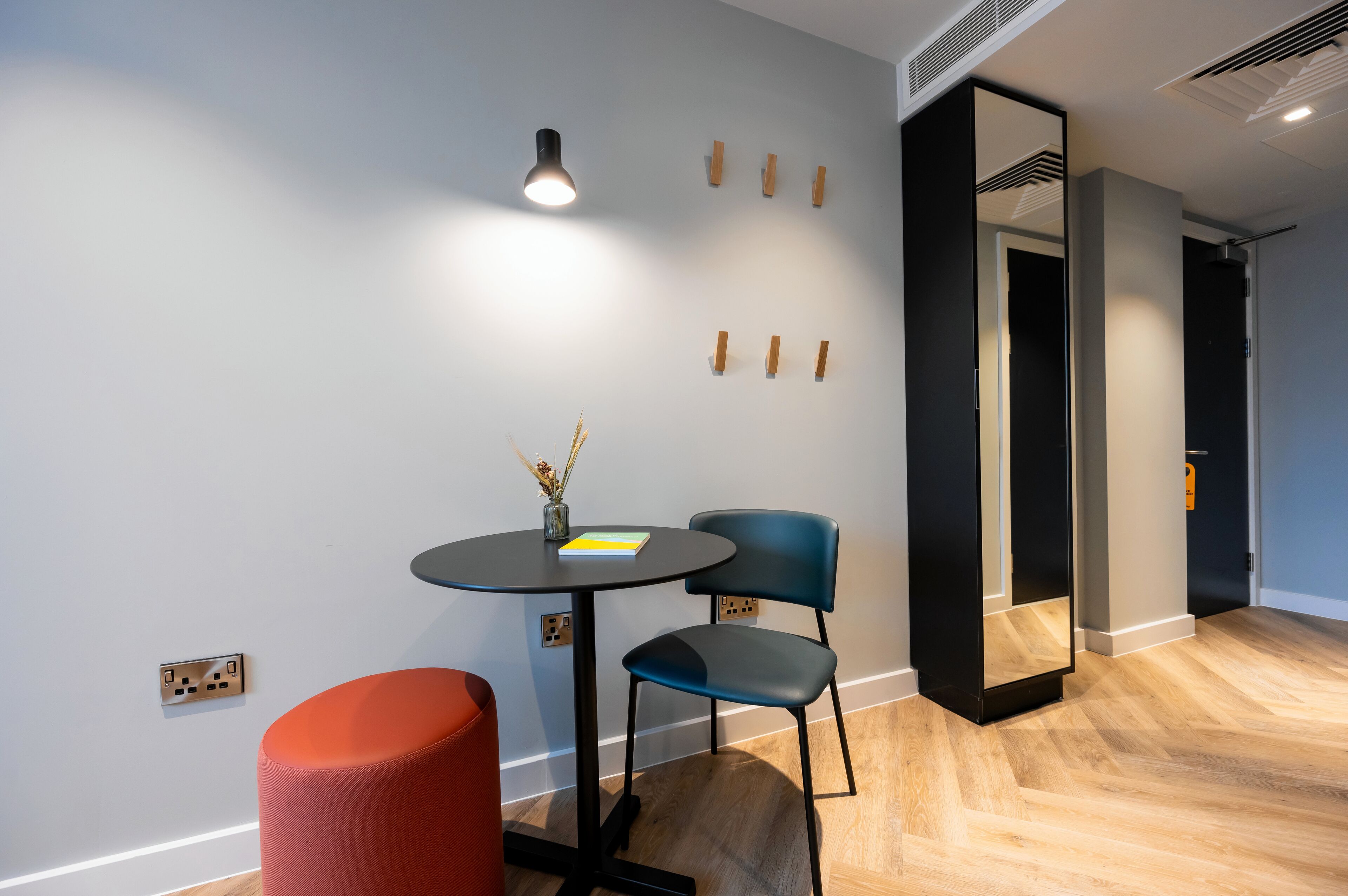 A modern room with a round table, two chairs, a red ottoman, a wall-mounted light, and a mirror.