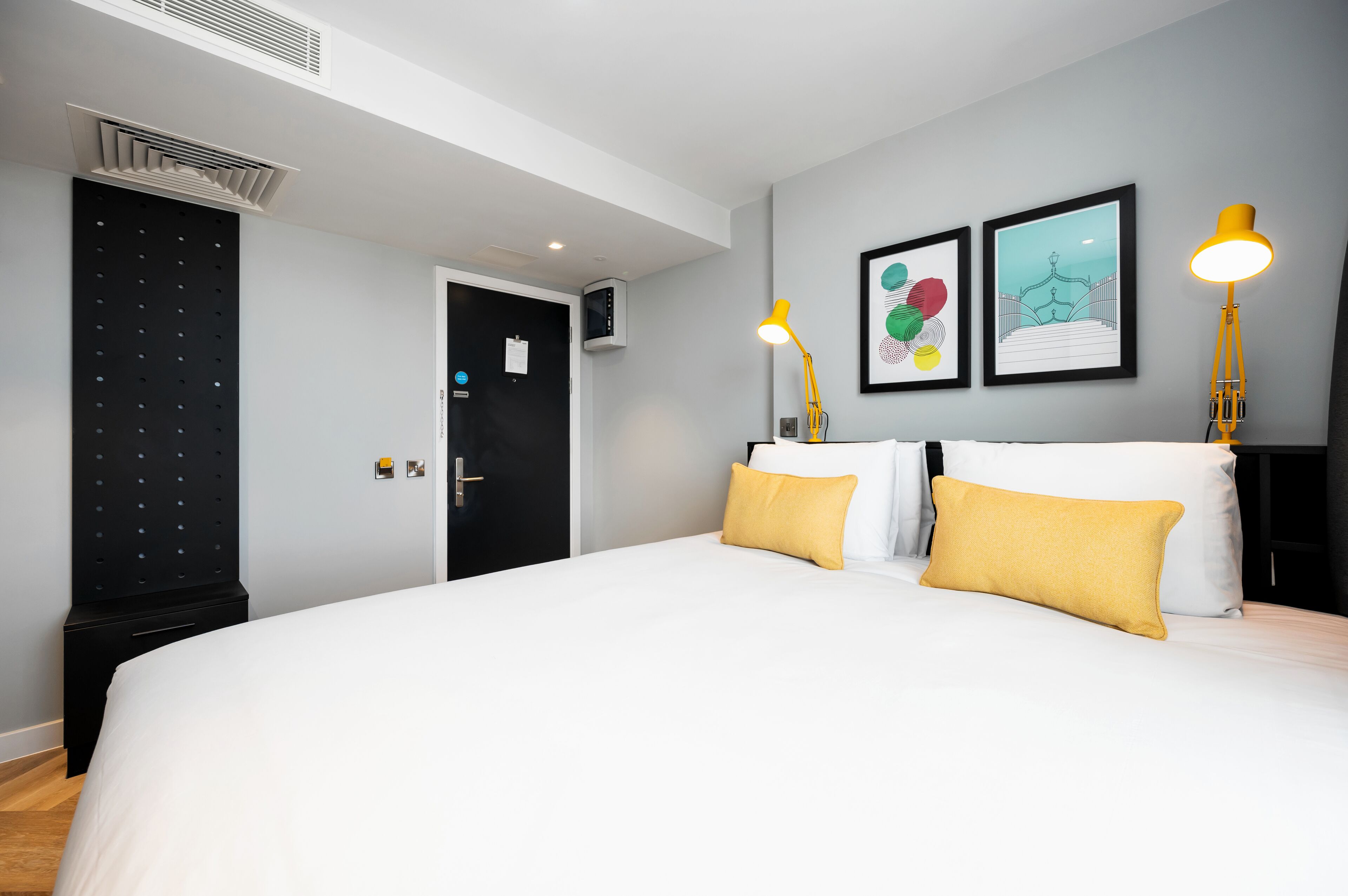 Habitación de hotel moderna con una cama grande, dos lámparas amarillas y obras de arte en la pared.