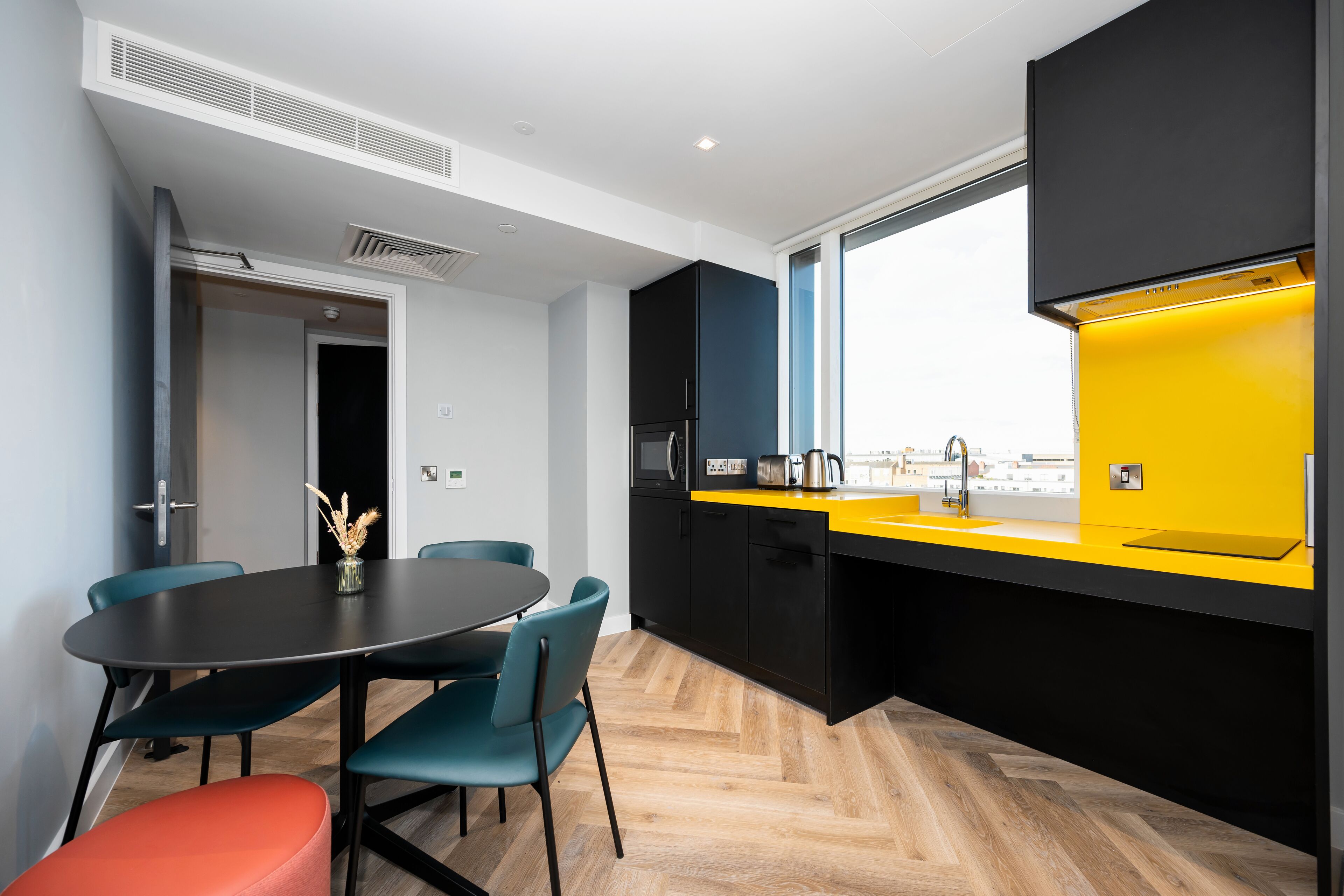 A modern kitchen with a black and yellow color scheme, a dining area with blue chairs, and a view through a large window.