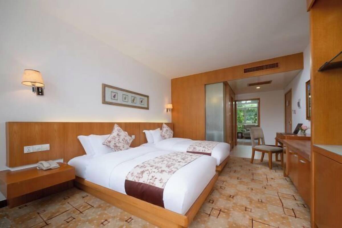 A hotel room with two beds, a wooden headboard, a bedside table with a phone, a framed picture on the wall, and a view of the outdoors through a window.