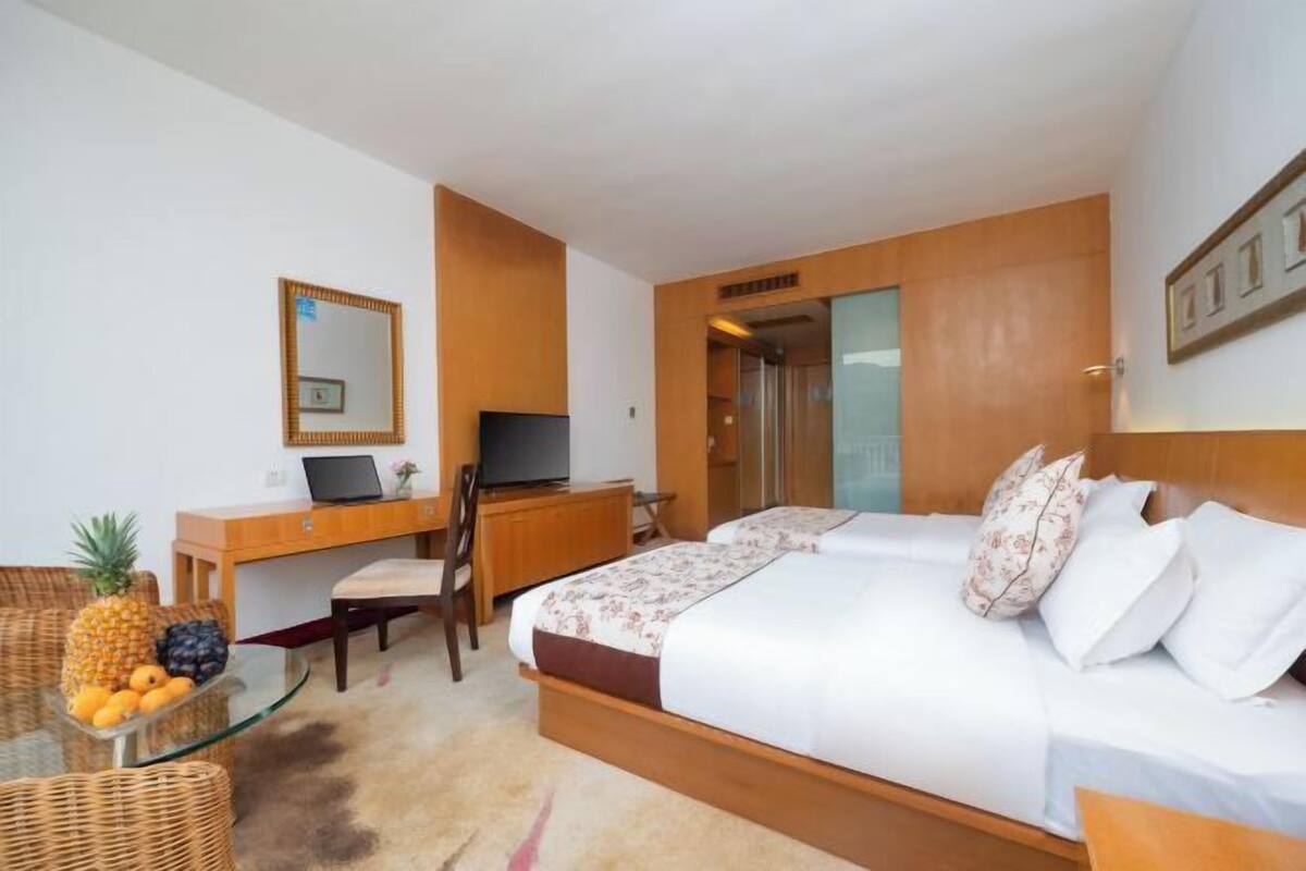 A hotel room with a bed, a desk with a computer, a chair, a small table with fruits, and a mirror on the wall.