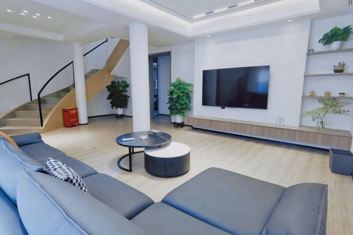 A modern living room with a sofa, a coffee table, a TV mounted on the wall, and a staircase.