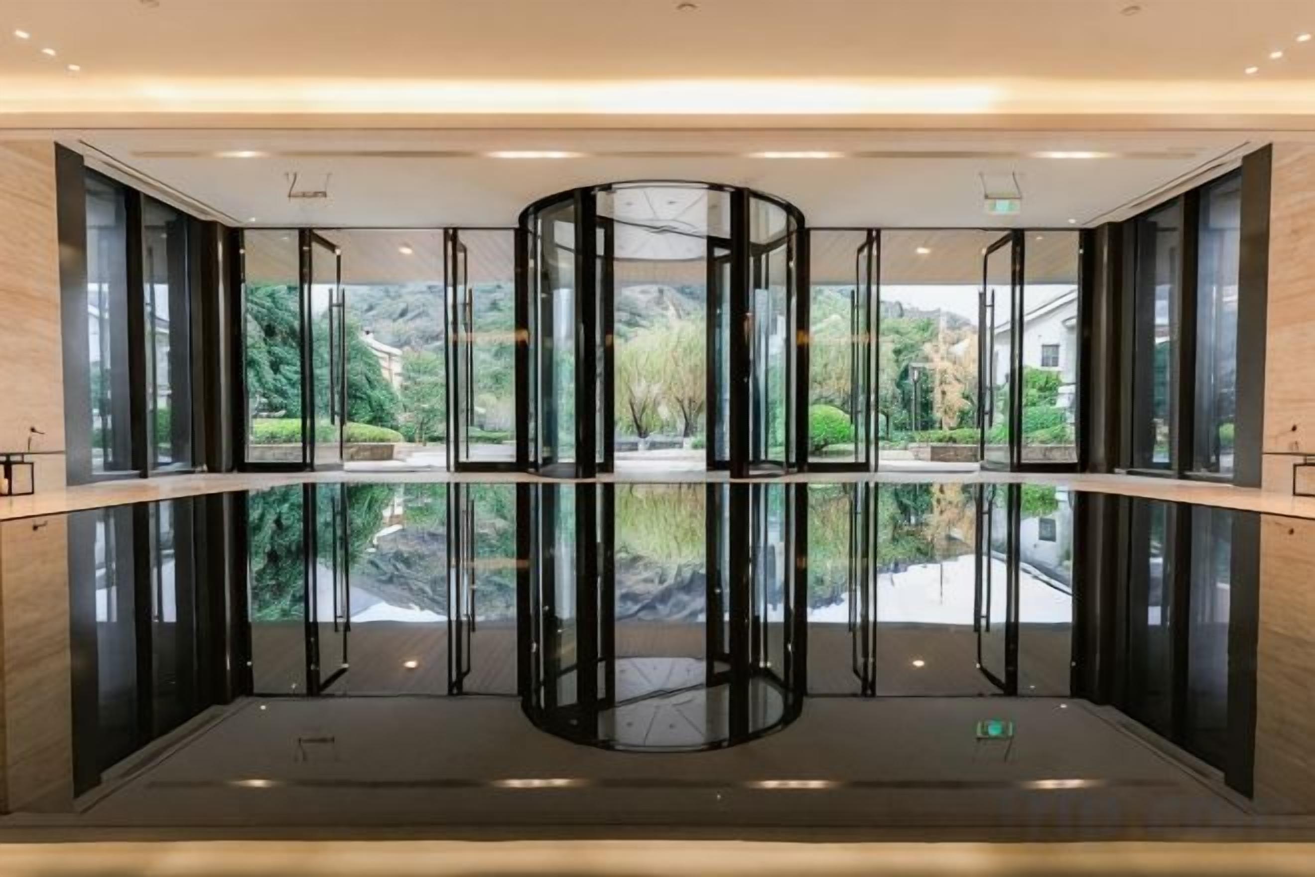 A revolving door with a glass enclosure, surrounded by glass walls, and a view of a garden outside.