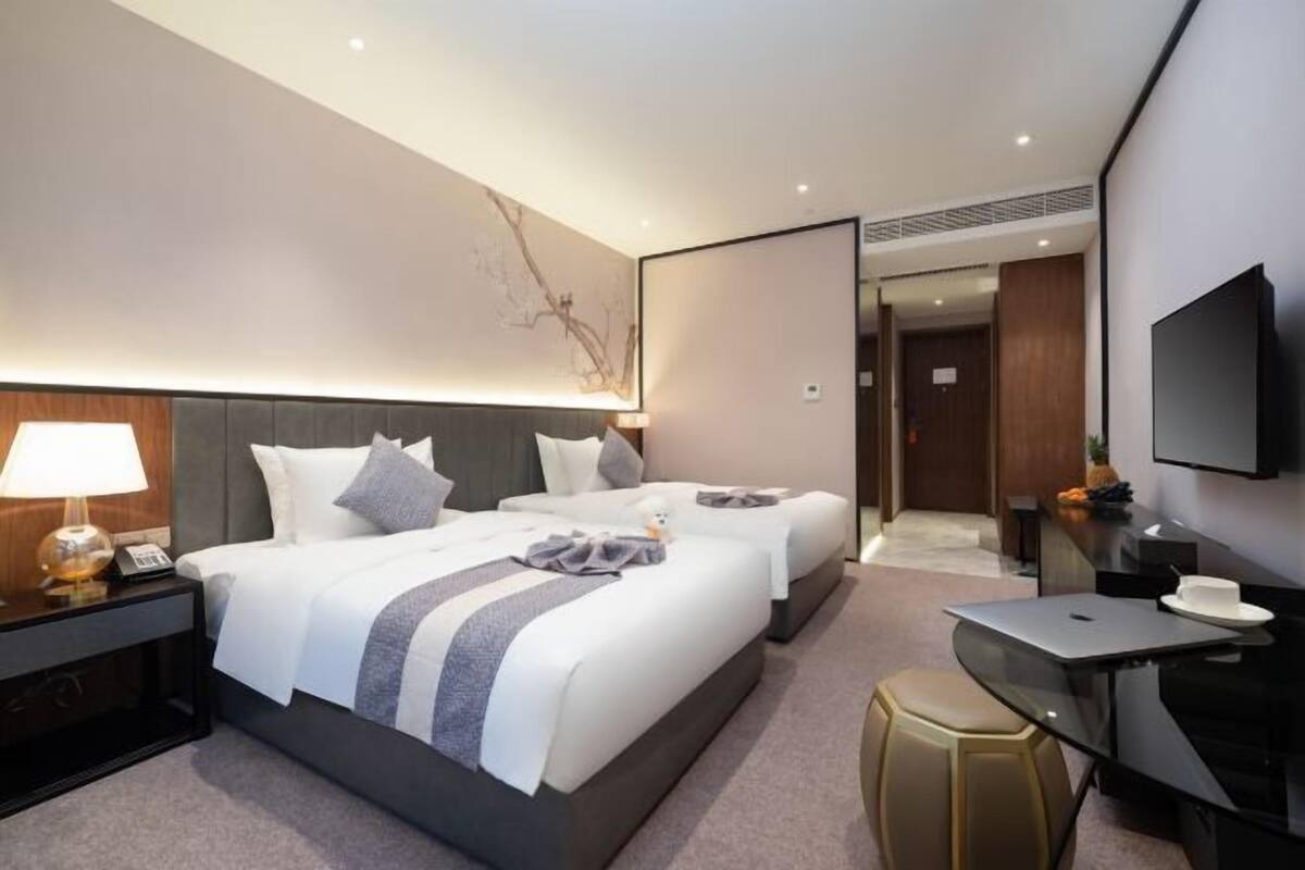 A modern hotel room with two beds, a desk, a chair, a TV, and a wall-mounted artwork.
