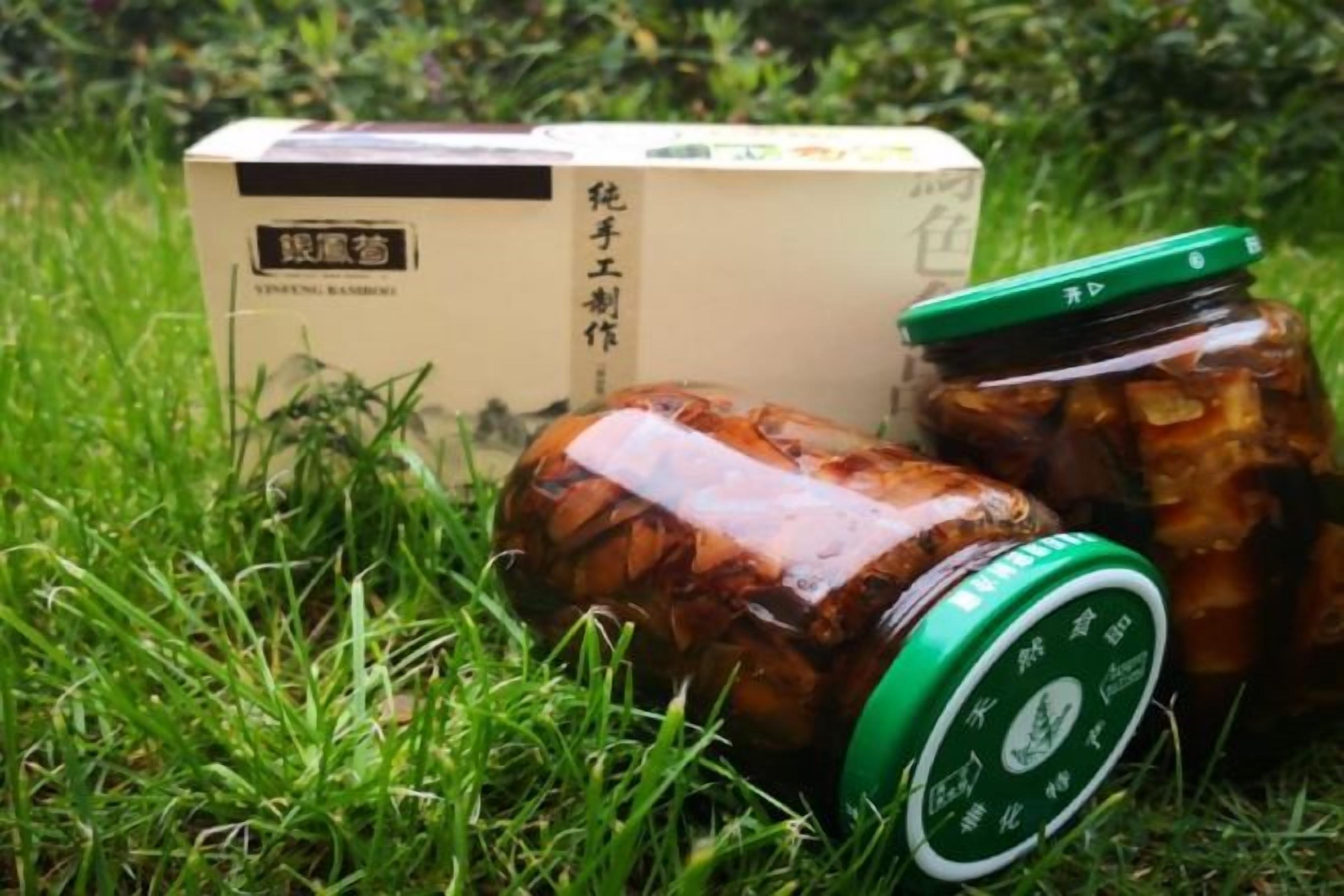 Une boîte étiquetée « prune vinaigrée » et un bocal avec un couvercle vert, posés sur l’herbe.